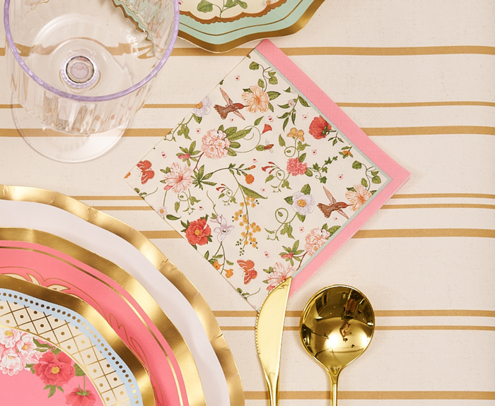 The Rococo Table Setting by Sophistiplate LLC features designer disposable plates in pink, gold, and white scallops, gold utensils, a clear glass, and a floral napkin with pink border on a cream and gold striped tablecloth.