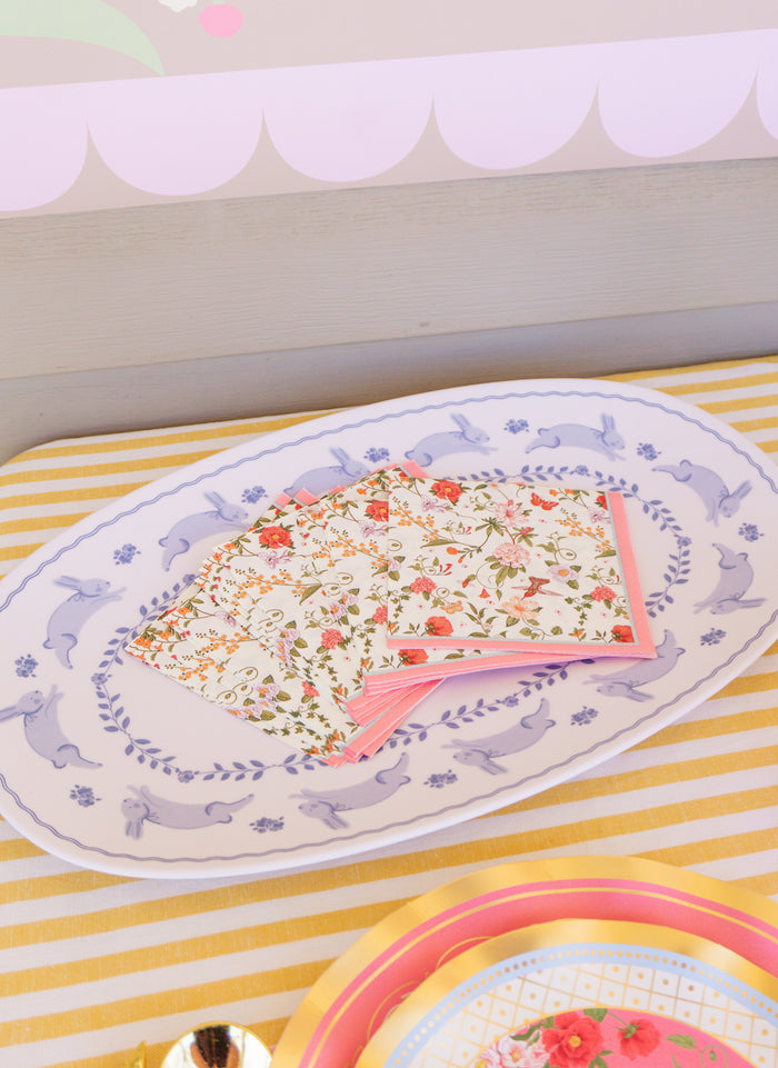 The Rococo Table Setting by Sophistiplate LLC features a white platter with a blue rabbit and leaf border, holding floral napkins with pink edges on a yellow-and-white striped tablecloth—ideal for your next party-in-a-box.