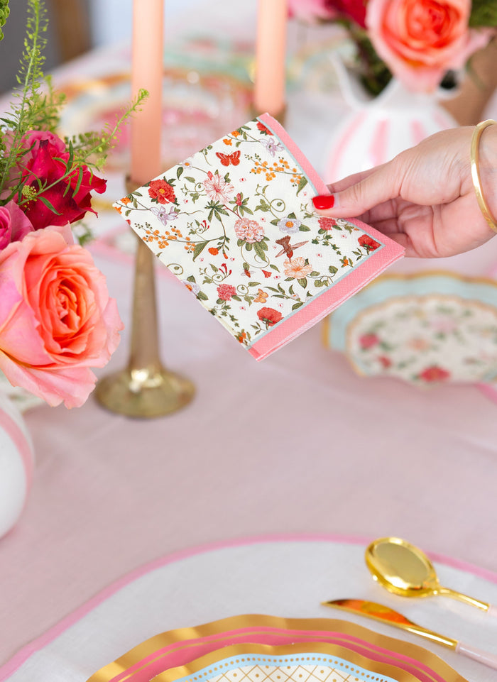 A hand with red nail polish holds a Sophistiplate Rococo Cocktail Napkin above a table set with gold cutlery, pink candles, vases of pink and orange roses, and chic pink and gold accented tableware.