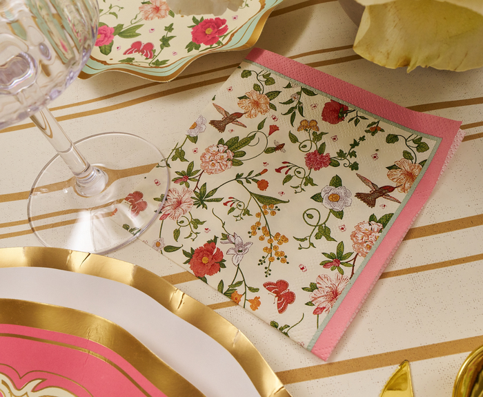 The Rococo Cocktail Napkins by Sophistiplate (26pk) add elegance to your table with colorful floral and bird designs, pink edges, and gold-trimmed decorative plates alongside a wine glass for a refined party look.