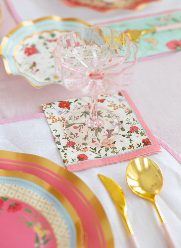 A pastel-themed table setting with a pink glass goblet, gold flatware, decorative plates with gold accents and florals atop a white tablecloth, and Sophistiplate Rococo Cocktail Napkins (26pk).