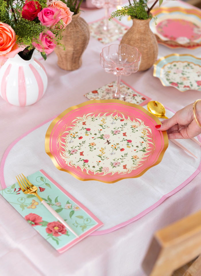 A hand sets a Rococo Table Setting by Sophistiplate LLC—a designer disposable plate with pink and gold floral accents—on a table adorned with matching napkins, gold utensils, colorful flower vases, and a light pink tablecloth.