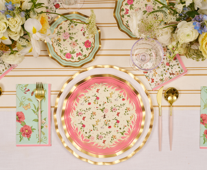 The Rococo Table Setting by Sophistiplate LLC features designer disposable plates with pink and gold trim, gold utensils, a floral napkin, glass, and lush flowers on a striped tablecloth—ideal for an elegant party-in-a-box.