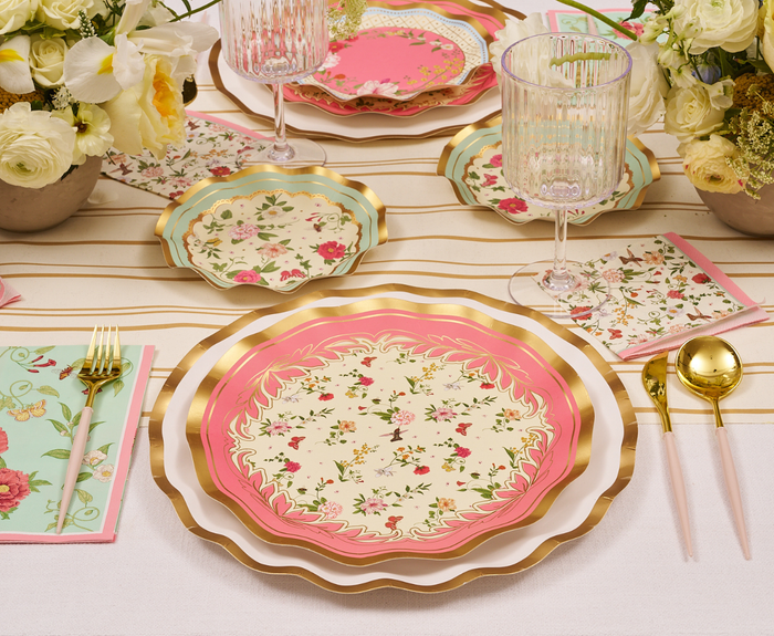 The Rococo Table Setting by Sophistiplate LLC features pink and gold designer plates, gold cutlery, clear ribbed glasses, floral napkins, and white and yellow flower decor for an elegant party-in-a-box tablescape.