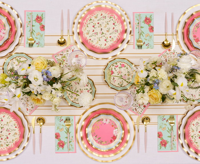 A decorative table setting with Sophistiplate Rococo Dinner Plates (8pk), gold utensils, pastel candles, rose napkins, and a centerpiece of white and yellow flowers with greenery on a light striped tablecloth.