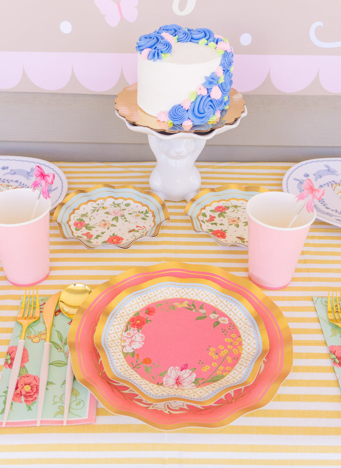 The Rococo Table Setting by Sophistiplate LLC features a pastel-themed party-in-a-box with floral designer plates, gold cutlery in floral napkins, pink cups with butterfly toppers, and a white cake with purple-blue frosting on a stand.