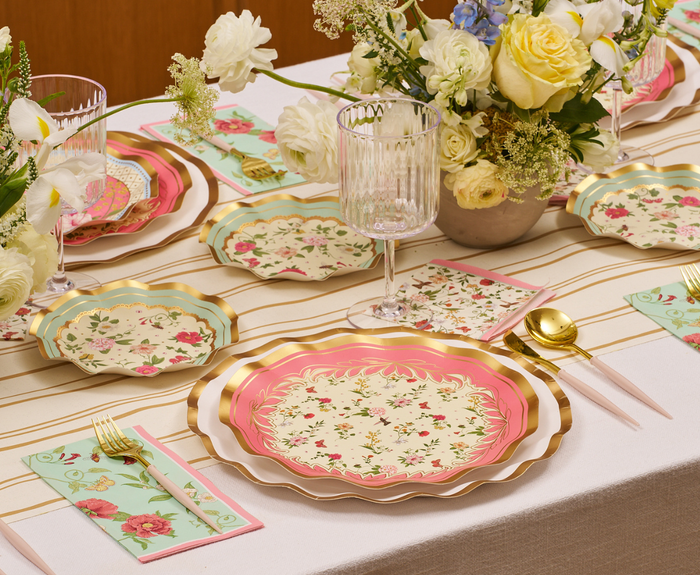 The Rococo Table Setting by Sophistiplate LLC showcases pastel floral designer disposable plates, matching napkins, gold cutlery, clear glassware, and white-yellow flower arrangements on a striped tablecloth for a refined dining display.