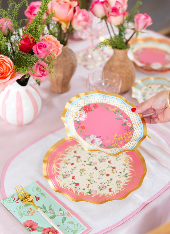 A hand sets a Rococo Table Setting by Sophistiplate LLC—pink and gold designer disposable plate, floral plates, gold cutlery, floral napkins, and rose vases atop a pink tablecloth—perfect for an elegant party-in-a-box experience.