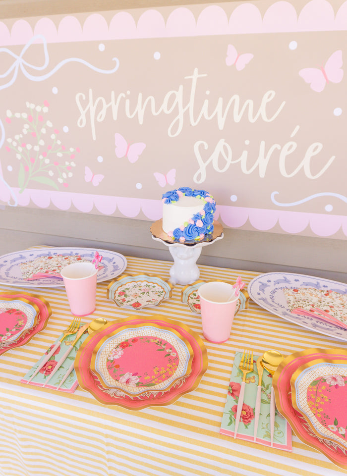 Create a dreamy spring soirée with the Rococo Table Setting by Sophistiplate LLC—designer disposable plates, pink cups, gold cutlery, and a small floral cake displayed against a pastel butterfly backdrop.