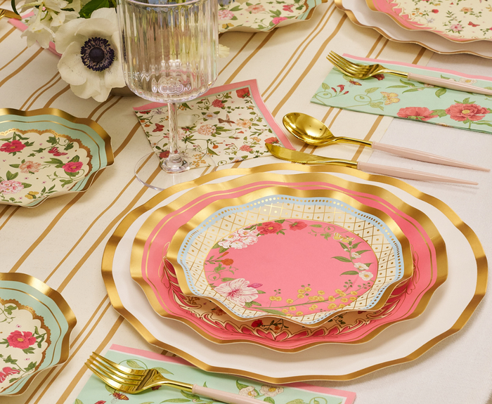 The Rococo Table Setting by Sophistiplate LLC features designer scalloped floral plates in pink, gold, and green, gold cutlery, a clear glass, and floral napkins on a cream tablecloth with gold stripes. White flowers accent the scene.