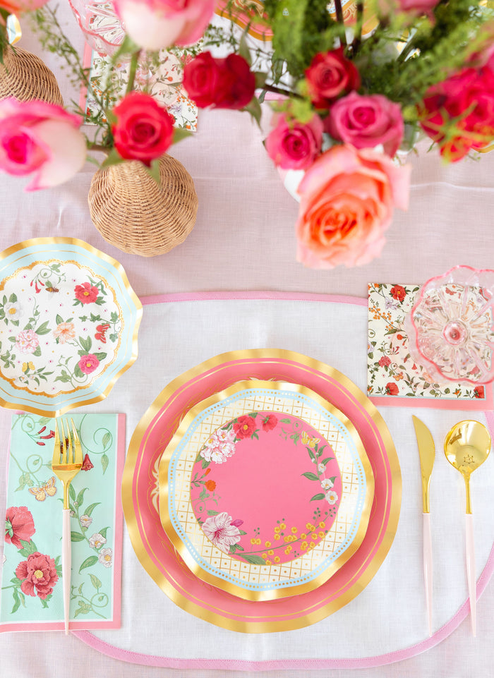 The Rococo Table Setting by Sophistiplate LLC features a designer pink and gold floral plate, gold utensils, a floral napkin, and a vivid pink-orange flower centerpiece on a light pink tablecloth—perfect for an elegant solo dining experience.