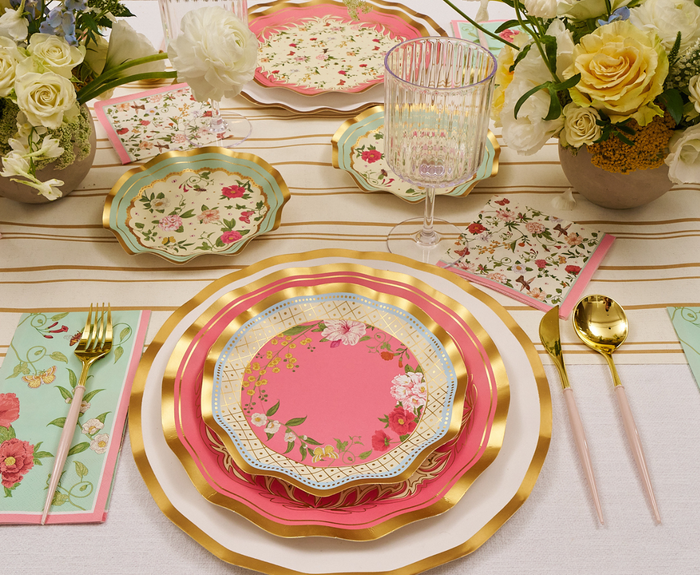 A table is set with Sophistiplate Rococo Salad Plates (8pk), floral napkins, pastel cutlery, a glass goblet, and vases of white and yellow flowers on a striped tablecloth.