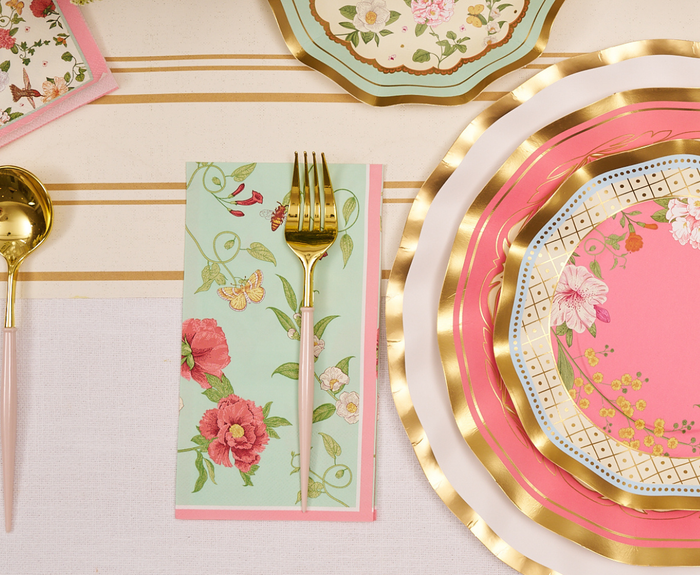 The Rococo Table Setting by Sophistiplate LLC features layered pink and gold designer disposable plates with floral designs, a gold fork and spoon, and a green floral napkin on a striped tablecloth.