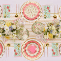 The Rococo Table Setting by Sophistiplate LLC features gold cutlery, designer pink and floral disposable plates, pastel floral napkins, and lush white, yellow, and blue flowers for an elegant tablescape on a white background.