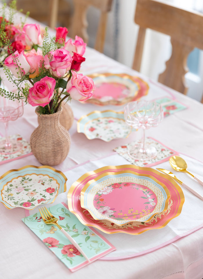 The Rococo Table Setting by Sophistiplate LLC features pink floral designer plates, gold utensils, floral napkins, ornate glasses, and a woven vase with pink roses on a light pink tablecloth—your perfect party-in-a-box.