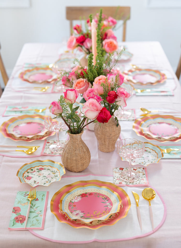 The Rococo Table Setting by Sophistiplate LLC includes pastel-themed designer disposable plates, gold cutlery, pink napkins, and pink glassware with a centerpiece of vases filled with pink and red roses—perfect for a stylish party-in-a-box.