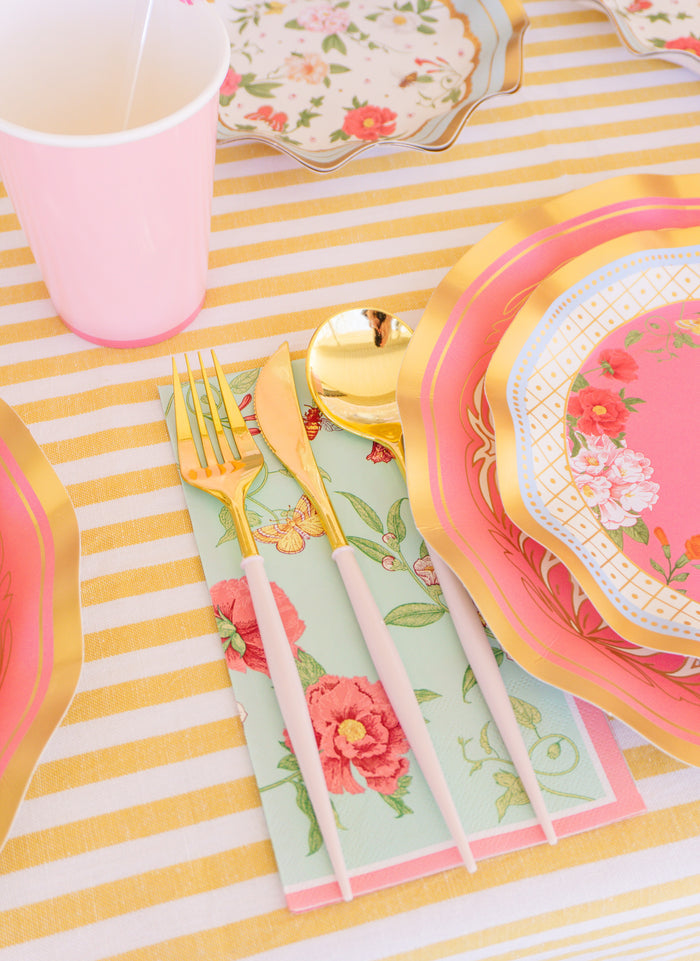 The Rococo Table Setting by Sophistiplate LLC features a pastel theme with a pink cup, gold and white cutlery on a floral napkin, designer disposable plates with gold trim, and a yellow and white striped tablecloth.