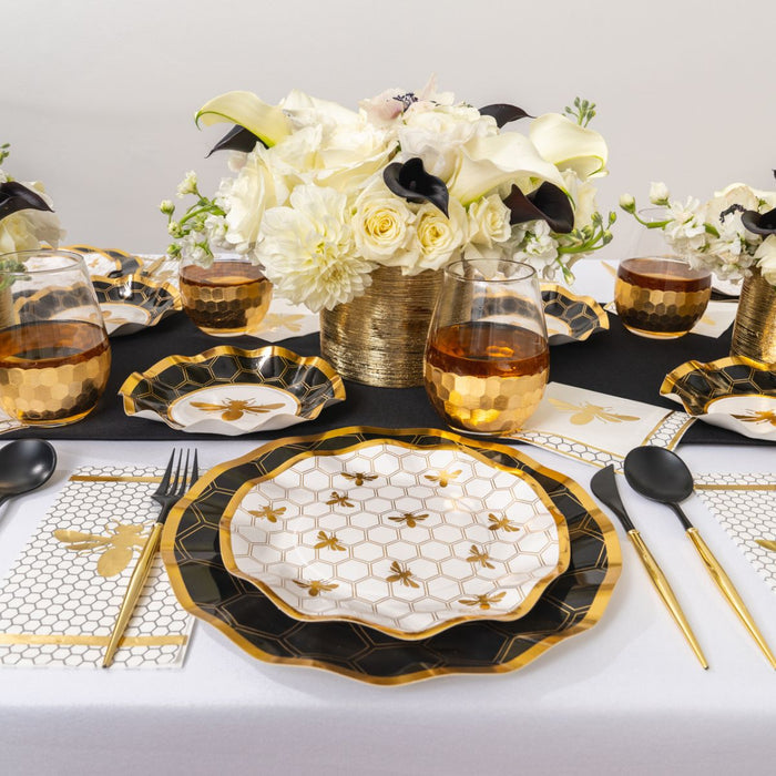 Create a stunning Rosh Hashanah table setting with the Sophistiplate Rosh Tablescape for 16 Guests: Honeybee, featuring elegant gold and black bee-themed heavy duty paper plates, matching black utensils, gold-rimmed glasses, and a beautiful centerpiece of white and yellow flowers in a gold vase.