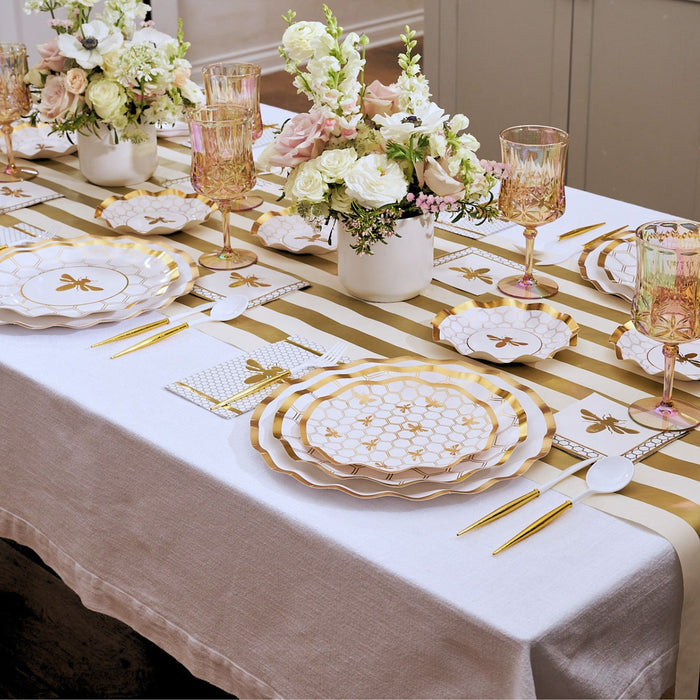 Introducing the White Honeybee Table Setting by Sophistiplate: an elegant arrangement featuring gold rim paper plates and disposable plates, gold utensils, a striped table runner, pink and gold glasses, and floral centerpieces with white and pale pink blooms in white vases—perfect for anyone seeking sophisticated party tableware.