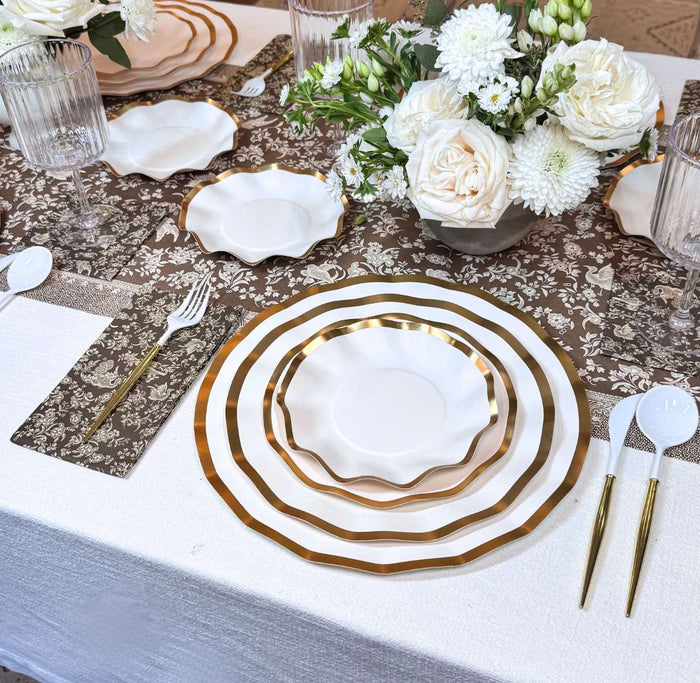 Create an elegant table with the Sophistiplate Royal Peacock Table Setting - Brown & White, featuring designer plates with gold rims, gold cutlery, clear glasses, and a white floral centerpiece on a brown and white patterned tablecloth.