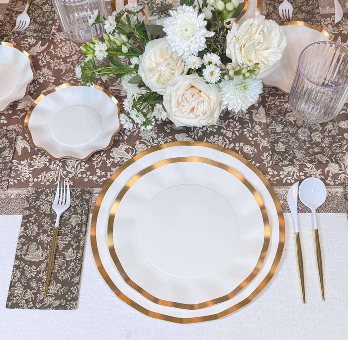 The Sophistiplate Royal Peacock Table Setting - Brown & White includes designer plates with gold trim, clear glasses, white cutlery with gold handles, brown patterned napkins, and a white and cream floral centerpiece on a brown tablecloth.