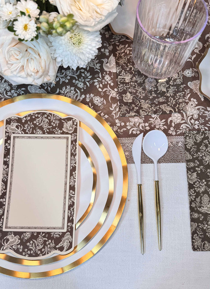 A close-up of the Sophistiplate Royal Peacock Table Setting - Brown & White shows designer plates, a floral menu card, white and gold cutlery, a clear ridged glass, and white flowers on a brown floral tablecloth.
