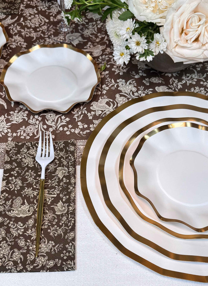 Create a stunning tablescape with the Sophistiplate Royal Peacock Table Setting - Brown & White: white plates with gold rims, gold-accented forks on brown floral napkins, and a centerpiece of white roses on a brown floral tablecloth.
