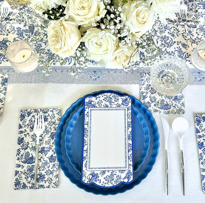 The Royal Peacock Table Setting - French Blue by Sophistiplate features blue and white floral napkins, designer plates, a menu card, silver cutlery, crystal glassware, white roses and baby's breath centerpiece, and a lit candle on a matching runner.