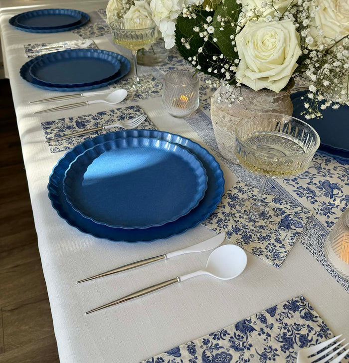 The Royal Peacock Table Setting - French Blue by Sophistiplate features designer party plates, white cutlery, patterned blue and white napkins, clear glassware, and a centerpiece of white roses and baby's breath on a white tablecloth.