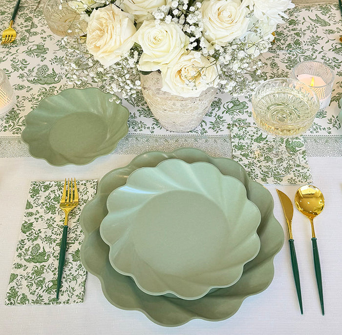 The Eco-friendly Table Setting- Sage by Sophistiplate LLC features green disposable plates, gold and green cutlery, a glass goblet, floral napkins, white roses with baby's breath in a vase centerpiece, and lit candles in the background.
