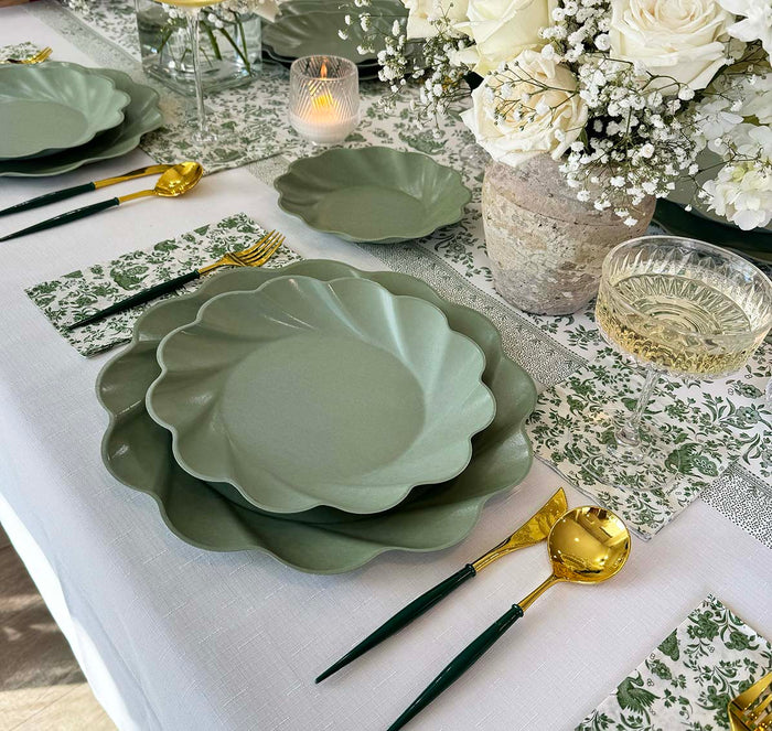 The Royal Peacock Table Setting - Sage by Sophistiplate features green scalloped disposable plates on a white tablecloth, gold and green cutlery, a floral-patterned napkin, a wine glass, candlelight, and a vase of white flowers with baby's breath.
