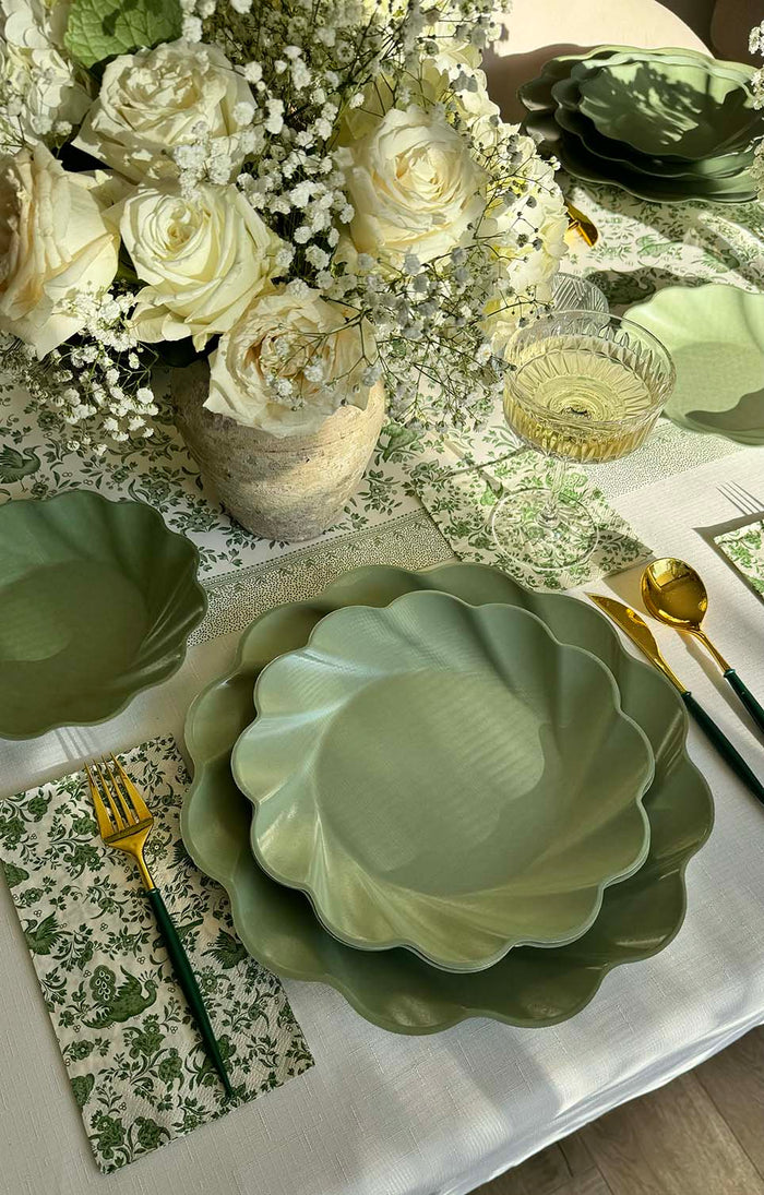 A table set with the Sophistiplate Royal Peacock Table Setting - Sage features designer disposable plates, gold cutlery with green handles, a floral napkin, glass of white wine, and a rustic vase of white roses and baby’s breath.