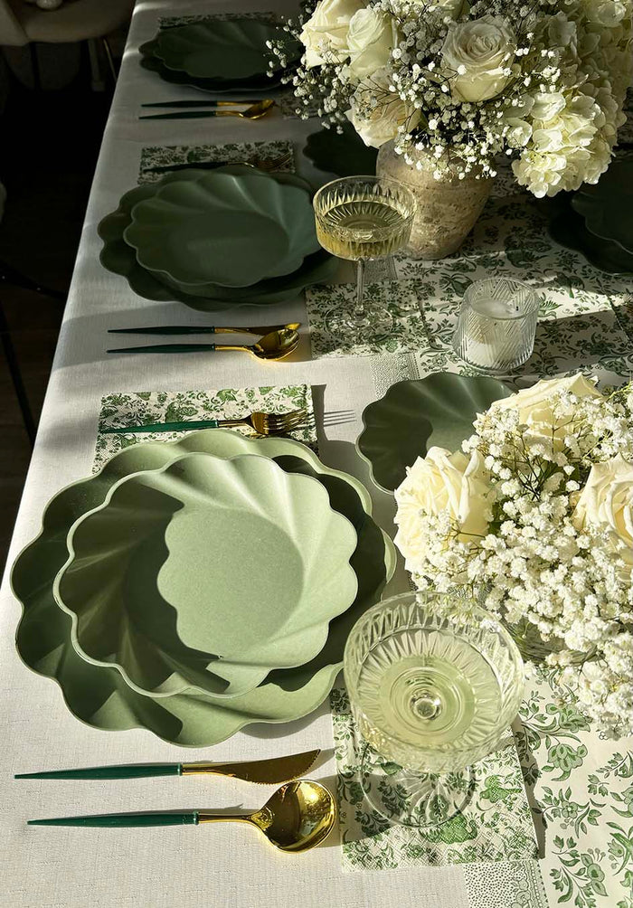 The Royal Peacock Table Setting - Sage by Sophistiplate features decorative green scallop plates, gold and green utensils, crystal glasses, green floral napkins, and a centerpiece of white roses with baby's breath.
