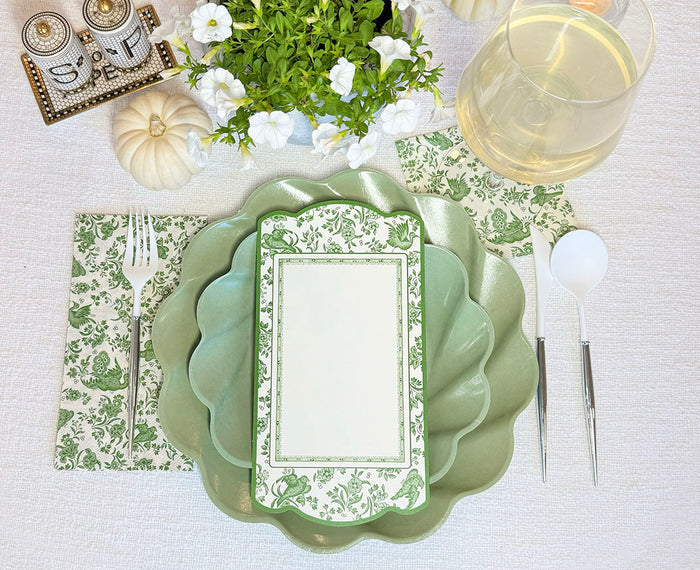 The Royal Peacock Table Setting - Sage by Sophistiplate features decorative green scalloped plates, floral napkins, white cutlery, a wine glass, mini pumpkins, a potted plant, and a salt & pepper set arranged on a crisp white tablecloth.