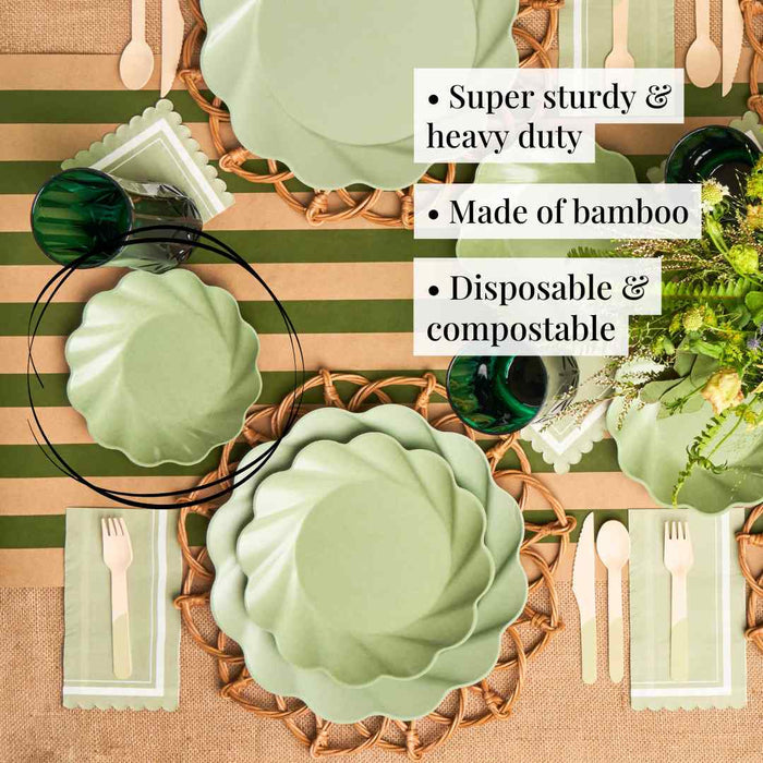 A table setting featuring Sophistiplate Simply Eco Salad Plate- Sage/8pk, wicker chargers, green glasses, wooden cutlery, a floral centerpiece, and a brown & green striped tablecloth. Super sturdy, eco-friendly, disposable & compostable plates.