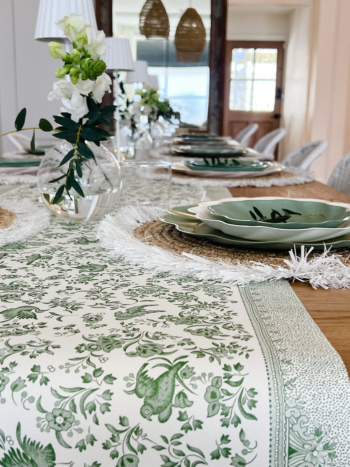 The Eco-friendly Table Setting- Sage by Sophistiplate LLC features disposable plates, woven placemats, a green-and-white floral runner, glass vases with white flowers, and wine glasses in a bright room.