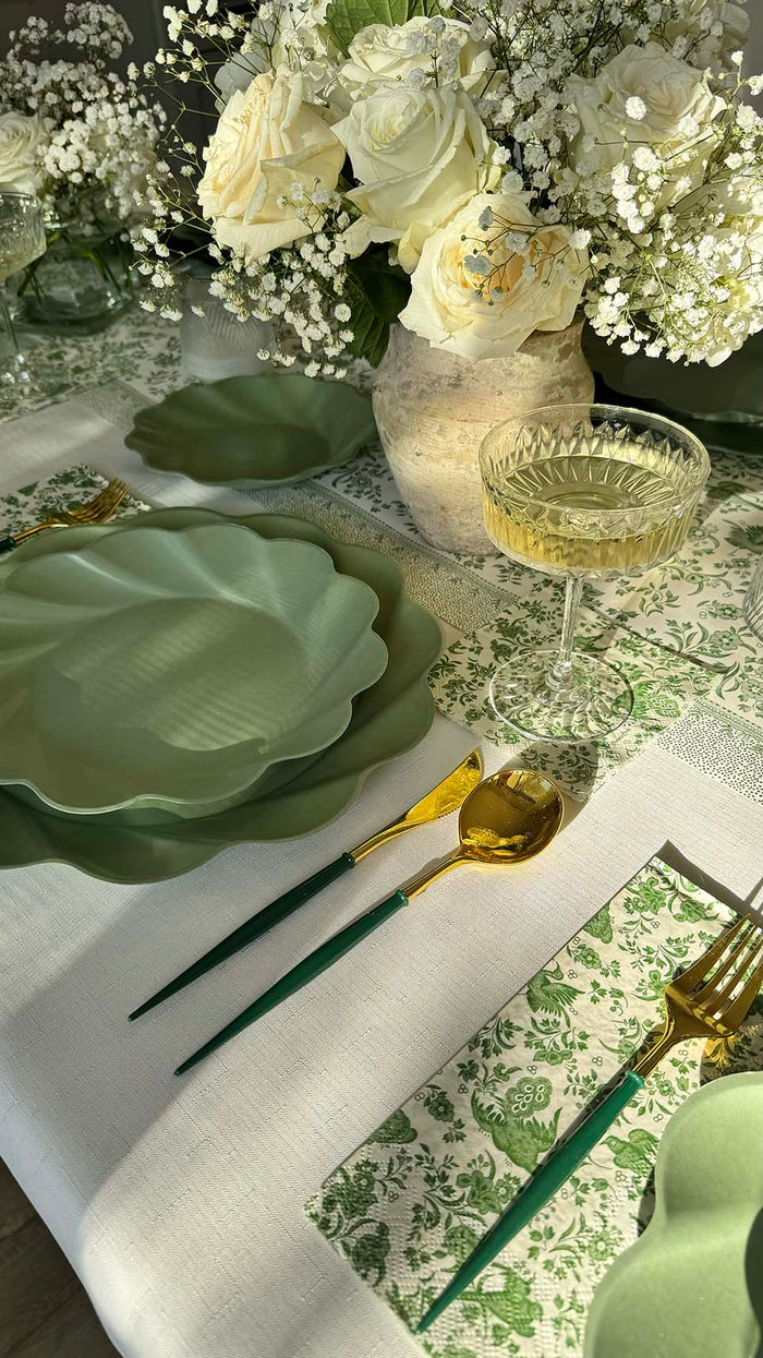 A close-up of the Sophistiplate Royal Peacock Table Setting - Sage, showcasing green leaf-shaped disposable plates, gold and green cutlery, a white wine glass, and a white floral centerpiece on a patterned green and white tablecloth.