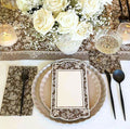 The Royal Peacock Table Setting - Sand by Sophistiplate features white roses in a vase, bamboo fiber plates, floral napkin with black cutlery, glassware, and a blank menu card on a brown and white floral-patterned tablecloth.