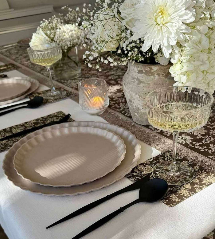 The Sophistiplate Royal Peacock Table Setting - Sand features cream bamboo fiber plates, black cutlery, patterned placemats, and glass goblets. A large vase with white chrysanthemums and baby's breath forms the centerpiece beside a lit candle.