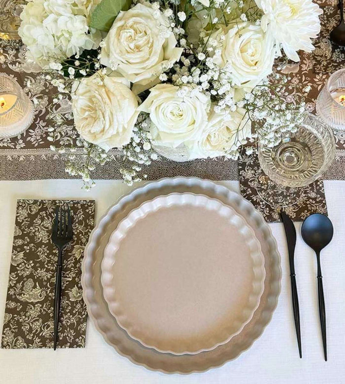 The Sophistiplate Royal Peacock Table Setting - Sand includes two round beige bamboo fiber plates, matte black cutlery, a floral napkin, a glass, and a white rose and baby's breath centerpiece on a decorated table runner.