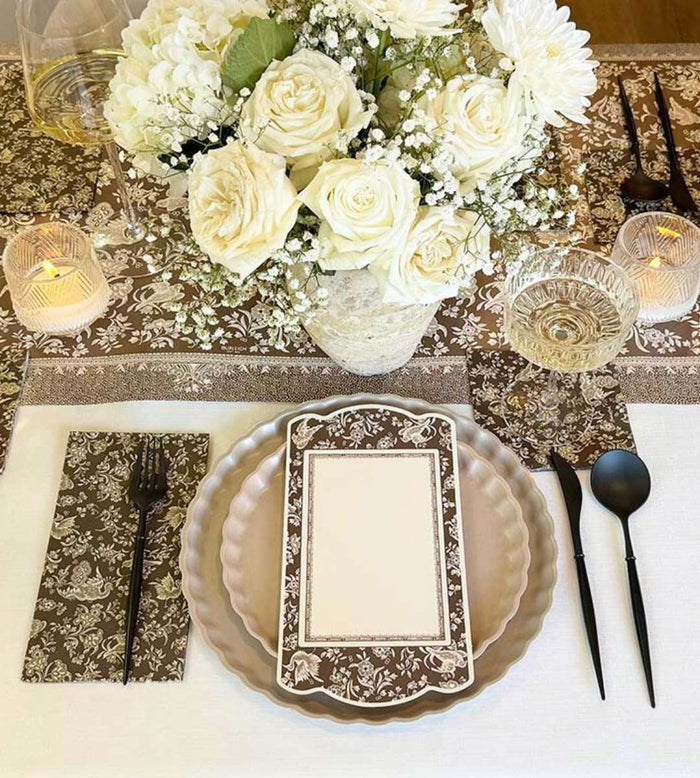 The Sophistiplate Royal Peacock Table Setting - Sand includes layered bamboo fiber plates, black cutlery on a patterned napkin, a decorative menu card, crystal glassware, candles, and a centerpiece of white roses and baby's breath.