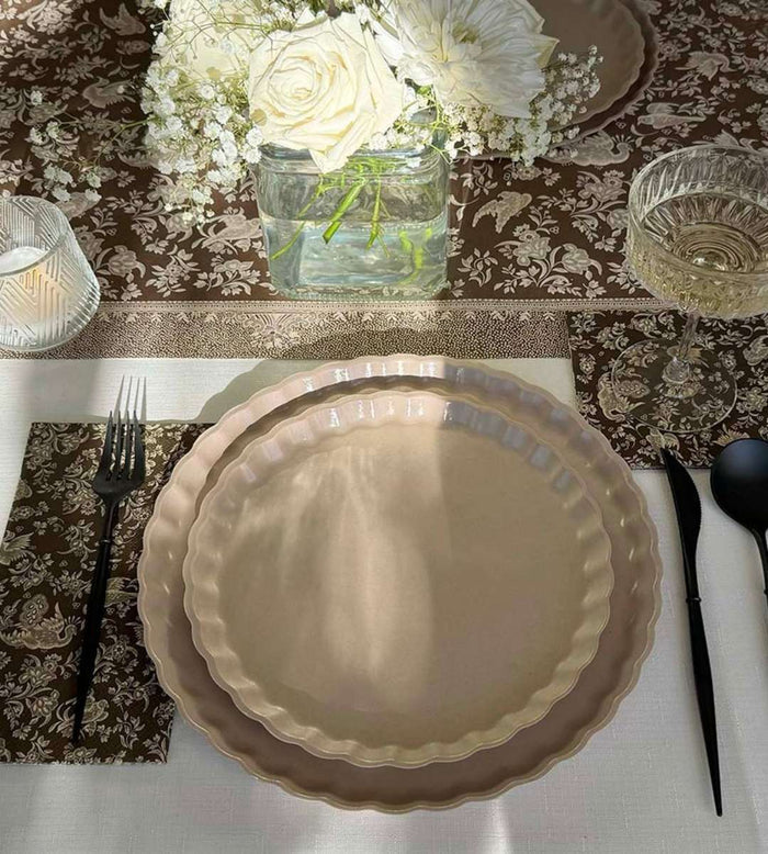 The Sophistiplate Royal Peacock Table Setting - Sand features two beige scalloped plates, black fork and spoon, wine glass, candle, and floral brown table runner with white flowers in a glass vase for an elegant tablescape-in-a-box look.