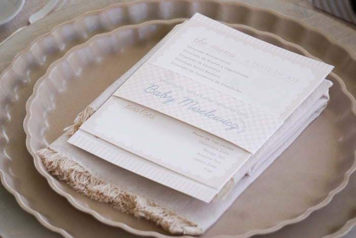 A cream menu card with elegant script sits on a fringed napkin atop the beige scalloped plate from the Eco-Friendly Table Setting- Sand by Sophistiplate, set for a formal meal with details like “A taste of joy” and “Baby Mislewicz”.