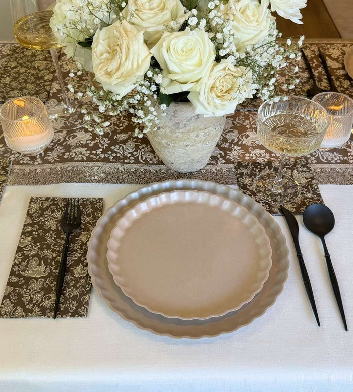 The Royal Peacock Table Setting - Sand by Sophistiplate features two stacked bamboo fiber plates, black flatware on a brown patterned napkin, a wine glass, candle, and a vase of white roses for an effortlessly elegant tablescape-in-a-box.