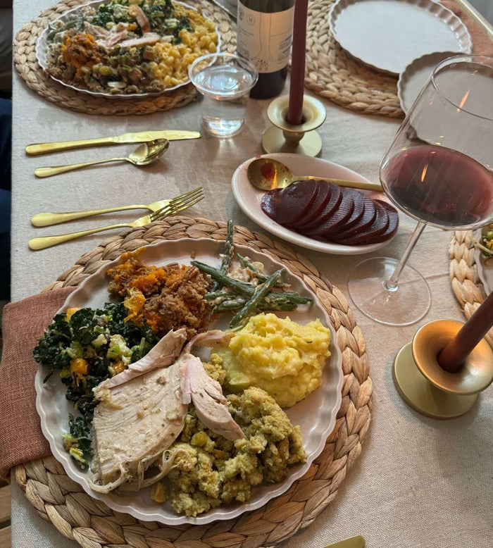 A meal is served on Sophistiplate's Eco-Friendly Table Setting- Sand, with bamboo plates, gold utensils, wine, candles, and woven placemats atop a beige tablecloth; turkey, sides, and salad complete the elegant look.