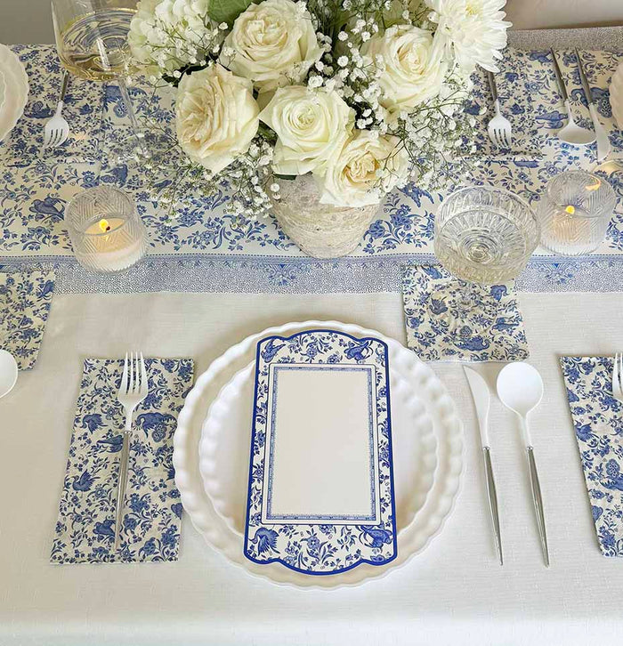The Royal Peacock Table Setting: Eco White & Blue by Sophistiplate features white and blue floral elements, bamboo fiber plates, a matching menu card, glassware, candles, and silverware—a perfect party-in-a-box for elegant gatherings.