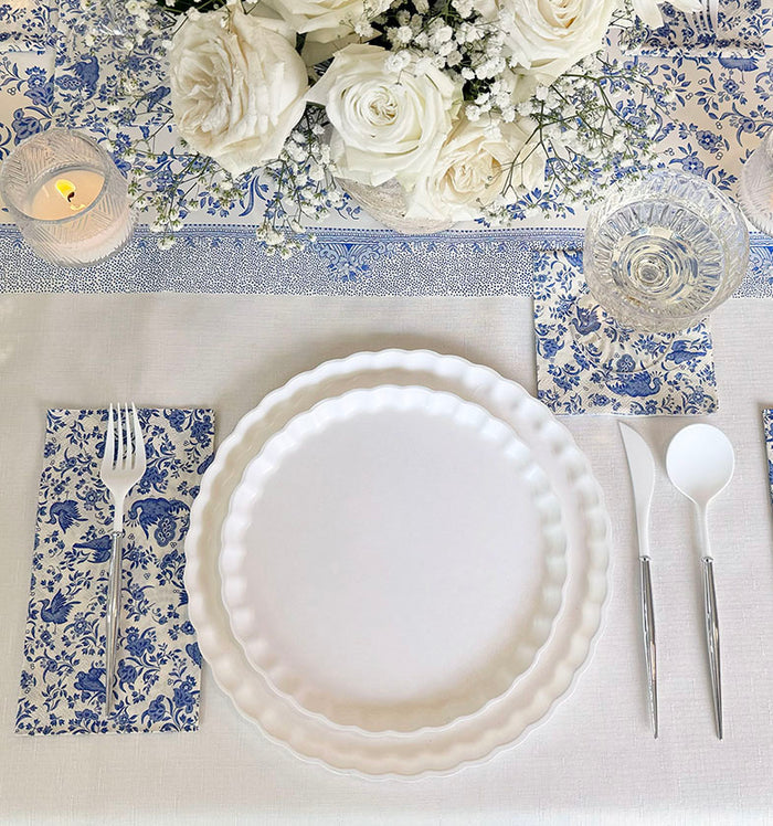 Create an elegant table with the Sophistiplate Royal Peacock Table Setting: Eco White & Blue—featuring white designer disposable plates, blue floral napkins, silverware, clear glassware, a tealight candle, and a centerpiece of white roses.