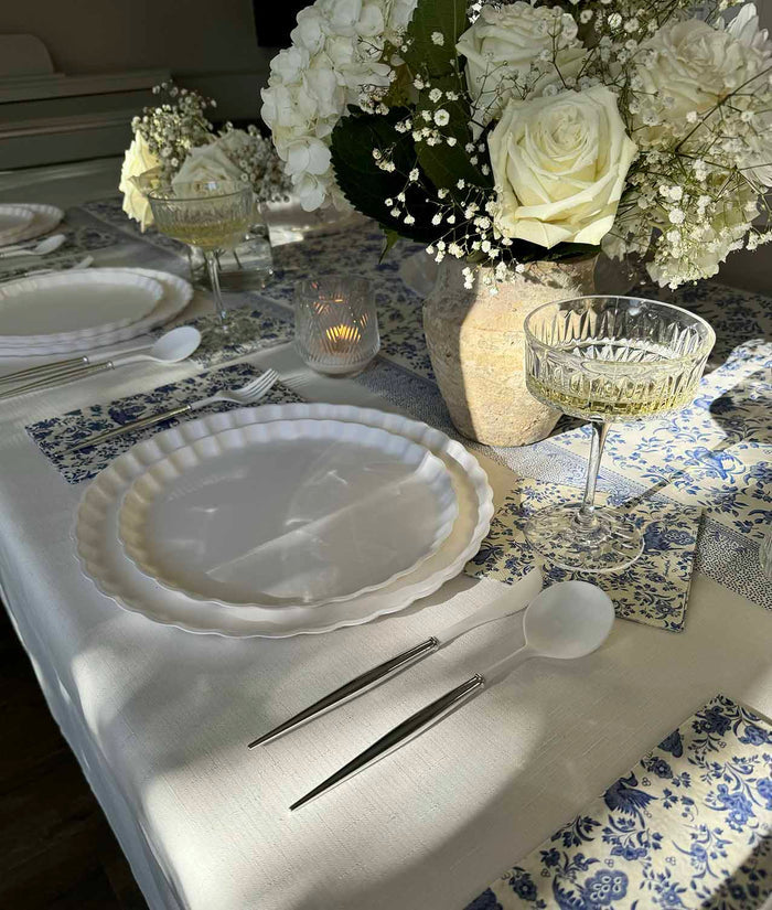 The Royal Peacock Table Setting: Eco White & Blue by Sophistiplate features bamboo fiber plates, silver cutlery, crystal glasses, and a centerpiece of white roses and hydrangeas—blue and white napkins complete this elegant party-in-a-box.