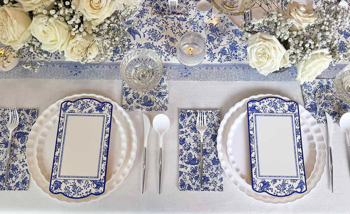 A close-up of the Sophistiplate Royal Peacock Table Setting: Eco White & Blue features blue and white designer disposable plates, matching napkins, white cutlery, and a floral centerpiece on a coordinated tablecloth.