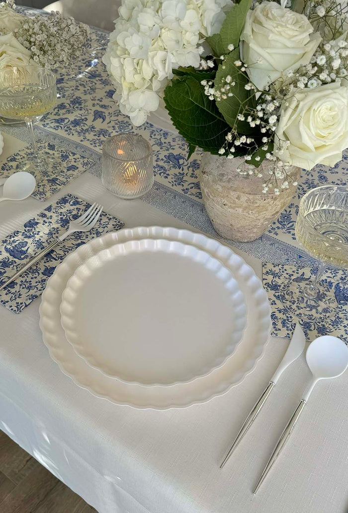A close-up of the Royal Peacock Table Setting: Eco White & Blue by Sophistiplate features designer disposable plates, silver cutlery, a blue and white floral napkin, glassware, and a vase of white blooms on a crisp white tablecloth.